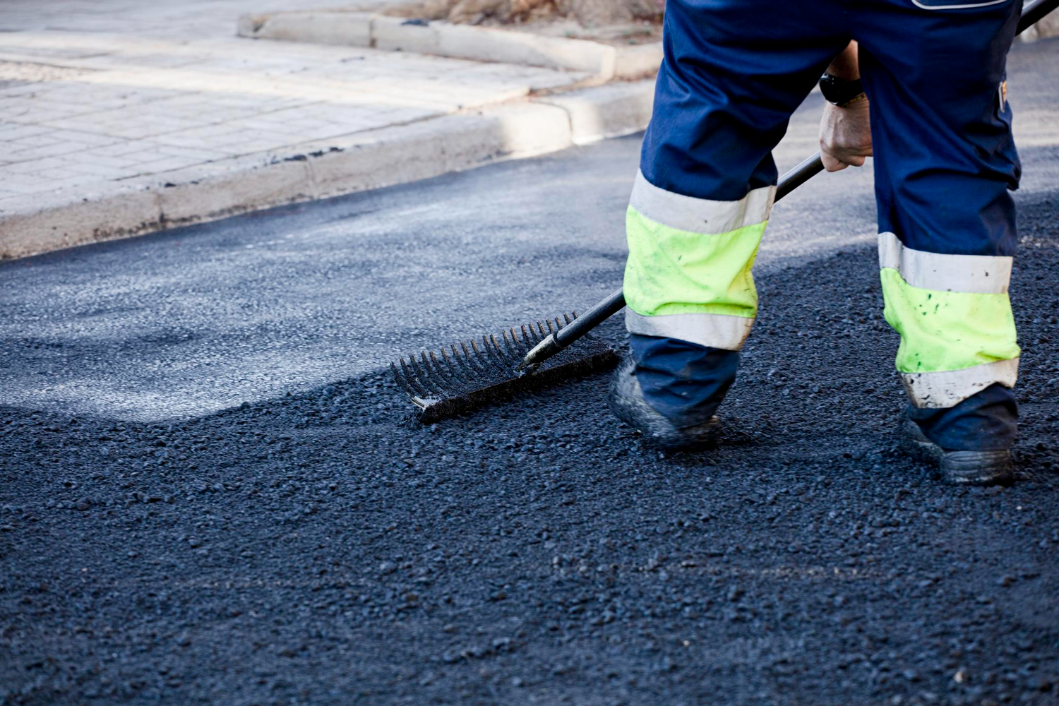 Worker spreading fresh asphalt on a road with a rake, wearing reflective pants and work boots.