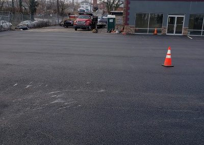 A single orange traffic cone stands in an empty, newly paved parking lot near a building and parked trucks—a testament to the quality work of J&S Paving, your trusted paving services provider in Edgewood MD.