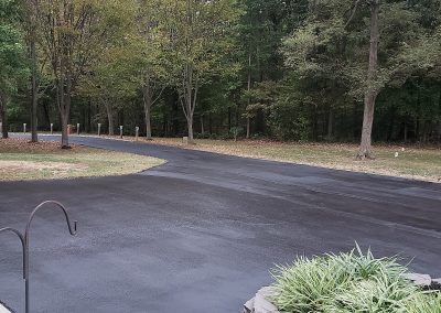 Freshly paved black asphalt driveway bordered by grass and trees in a wooded residential area, expertly completed by J&S Paving. Serving Edgewood MD with top-quality paving services.