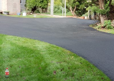 Freshly paved black asphalt driveway curves past a house with green lawn and trees in Edgewood MD; soda bottle on grass—a great example of quality Paving Services in Maryland.