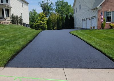 A freshly paved black asphalt driveway by J&S Paving in Edgewood, MD stretches between two houses, bordered by neatly trimmed green grass.