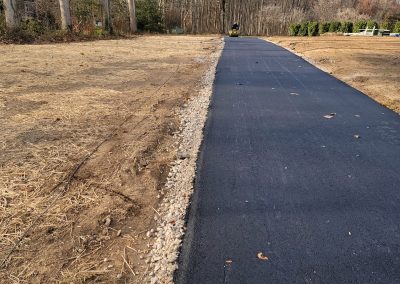 Newly paved asphalt path with gravel edges by J&S Paving, running through a mostly barren, grassy area with trees in the distance.
