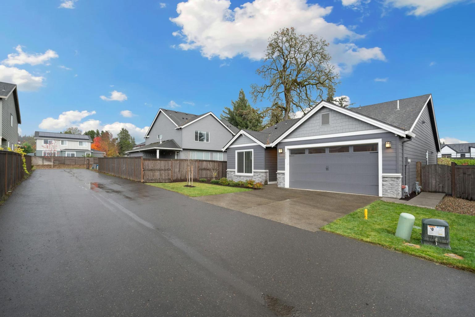 Single-story gray house with a two-car garage on a wet driveway, under a blue sky with clouds.