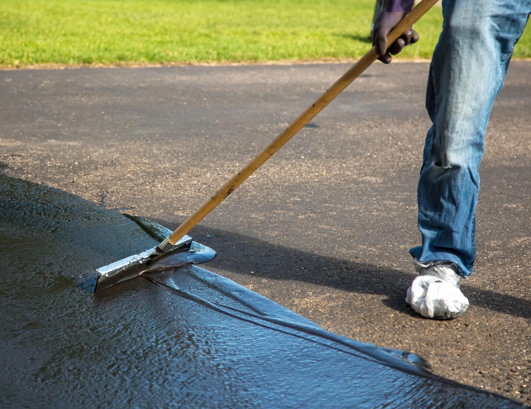 sealcoating-img Person spreading black sealant on driveway with a long-handled tool, grass visible in the background.