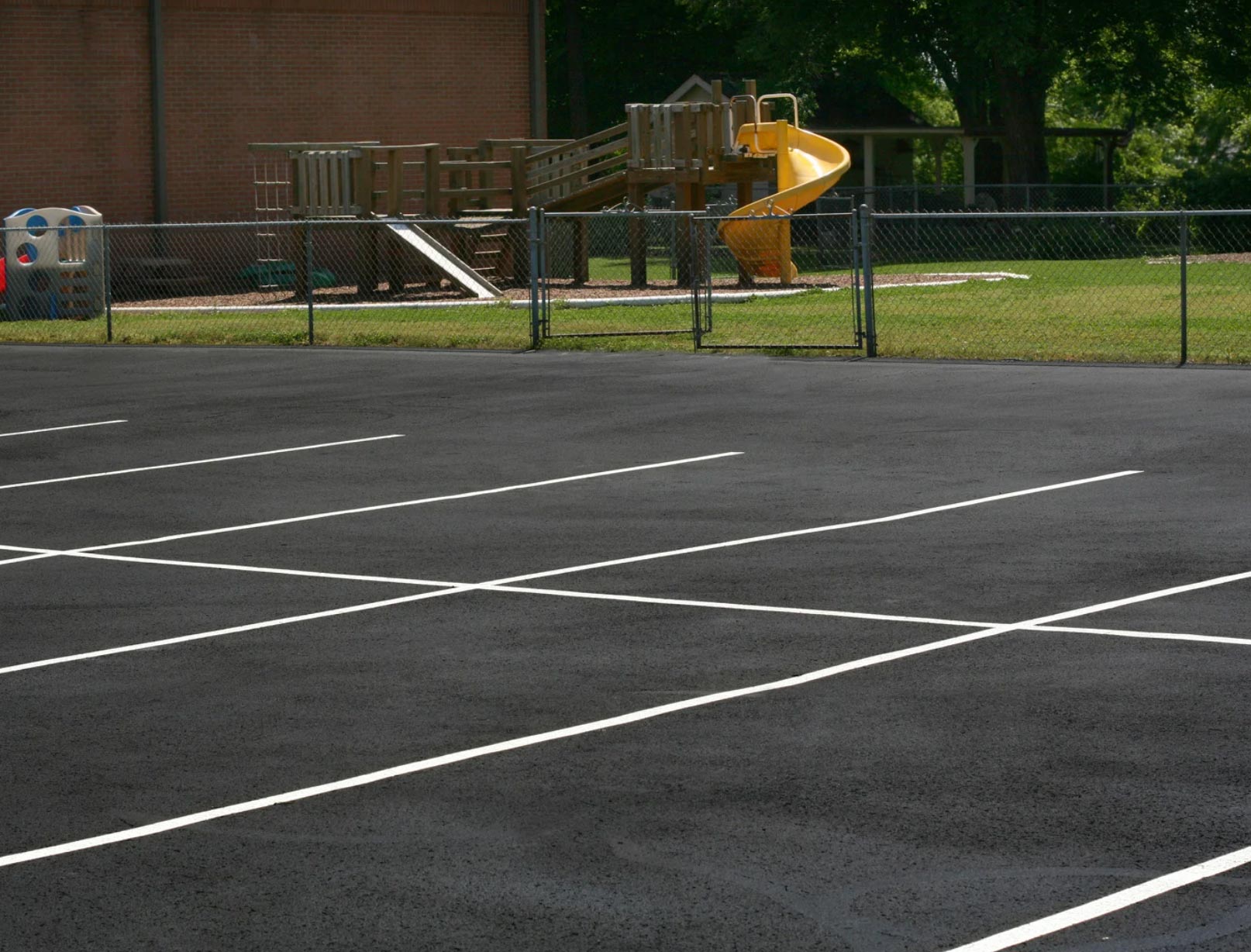 parking-lot-img-2 Empty asphalt parking lot with white lines, a fence, and a playground with a yellow slide in the background.
